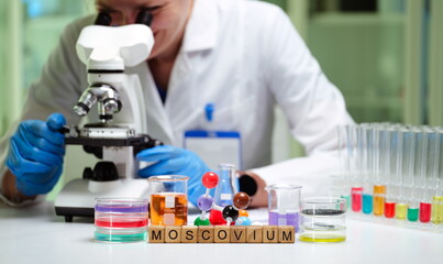 A dedicated scientist uses a microscope, surrounded by colorful chemical samples labeled MOSCOVIUM.