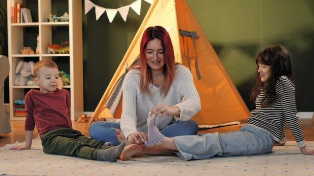 Mother tickling daughter's feet while son is sitting on carpet in playroom