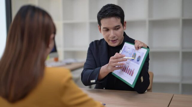A man is presenting a piece of paper to a woman, pointing to the numbers on it. The numbers appear to be financial data, and the man is likely explaining the information to the woman