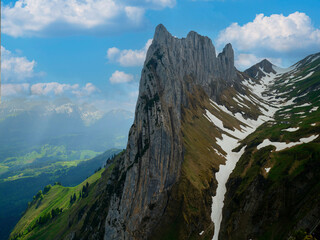 Majestic mountain peak rises above verdant hills and patches of snow under a bright blue sky filled with fluffy clouds