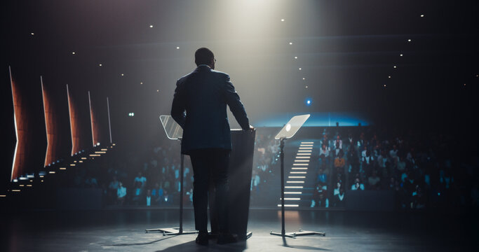 Business Leader Walking Out on Stage to Give a Talk at a Global Finance Summit. Audience Listening to His Insightful Talk. Speaker Standing with His Back to Camera While Making a Motivational Speech