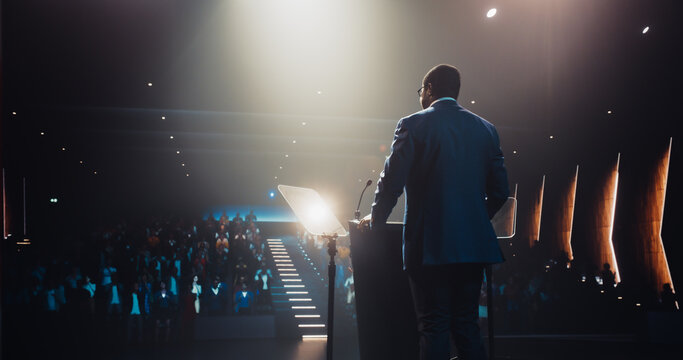 Confident Thought Leader Walking to a Tribune with laptop Computer, Delivering an Inspiring Presentation About Innovation at Technology Conference. Black Man Talking to an Audience in a Dark Venue - Powered by Adobe
