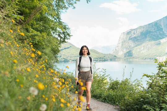 Glowing with joy, a woman strolls a nature trail wrapped in greenery, grinning as mountains and a sparkling lake unfold her perfect summer reset