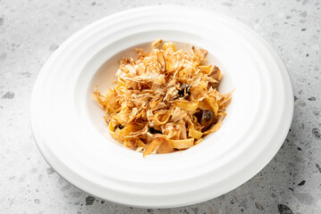 A white plate with pasta, topped with delicate bonito flakes, served on a speckled gray countertop....