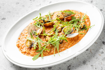 A plate of creamy tomato risotto garnished with fresh arugula and clams, served on a white dish. The gourmet seafood meal is presented on a speckled gray surface.