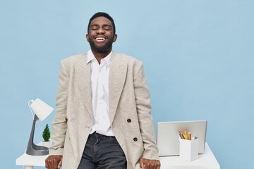 Smiling man in beige blazer and white shirt standing beside modern office desk with laptop,...