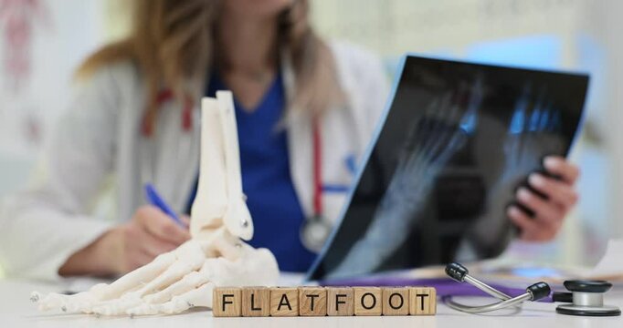 Doctor holds examining X-ray picture diagnosing Flatfoot shown on wooden blocks. Anatomical model and medical equipment suggests clinical workplace