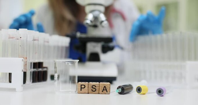 Letters PSA on wooden blocks with blood samples in test tubes on desk. Lab assistant conducts bio materials analysis with microscope in clinic