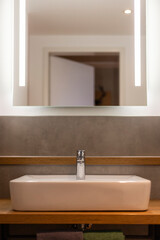 A sleek bathroom sink is placed on a wooden countertop, paired with a minimalistic illuminated mirror in a modern space. Soft lighting adds to the ambiance of the room