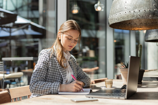 Young businesswoman writing in notebook while working at laptop in sunny outdoor café. Stylish woman in blazer focused on planning or studying with coffee and cactus nearby