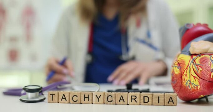 Word Tachycardia on wooden blocks with heart model against doctor at desk. Female cardiologist diagnoses cardiac disease at appointment in hospital