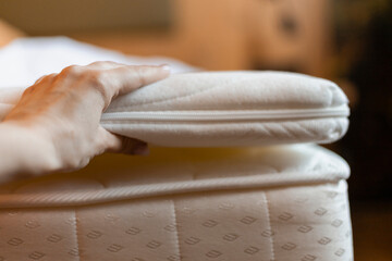 A hand gently lifts a comfort layer from a mattress in a softly lit bedroom. The bedding appears...