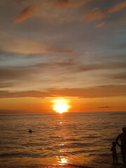 Fototapeta premium Seascape photography. Thick horizontal clouds. Tropical sky in orange colors. Panorama of the southern sea. Sun path in the water. Sunset ocean landscape for wallpaper. Human silhouettes on the shore.