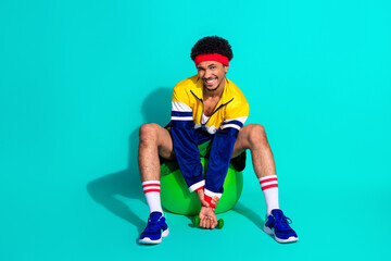 Energetic young man in retro sportswear smiling on a teal background while sitting on an exercise ball and holding a dumbbell