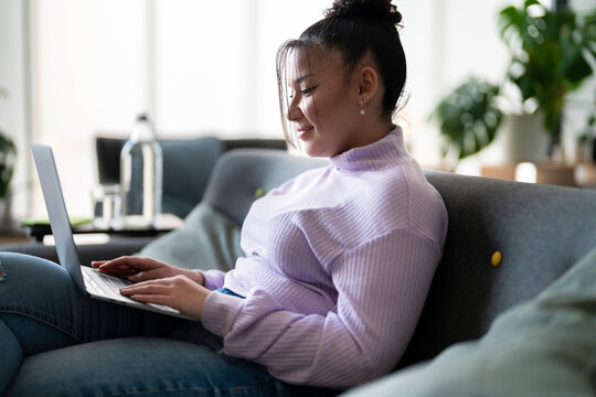 Young woman sits comfortably on a sofa at home, focused on her laptop screen. She is working or studying remotely in a relaxed environment.