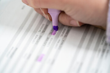 A person's hand uses a purple highlighter pen to mark specific numbers on a printed document. This process involves reviewing and emphasizing key figures within a financial report or data sheet.