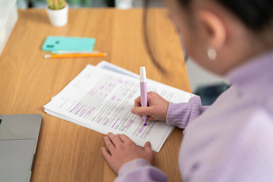 A young person highlights text on a document with a purple marker while reviewing papers at a wooden desk. This person is focused on their work.