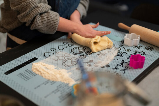 Close up of hands kneading dough on a patterned baking mat. Flour, a rolling pin, and cookie cutters are nearby. Preparing for baking cookies or pastries.