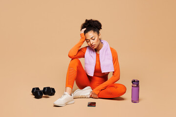 Full body young sad exhausted ill fitness trainer instructor sporty woman sportsman wear orange clothes towel spend time in gym sit isolated on plain beige background. Workout sport fit abs concept.