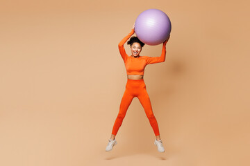 Full body young happy fitness trainer instructor sporty woman sportsman she wear orange clothes spend time in gym jump high with fit ball isolated on plain beige background. Workout sport abs concept.