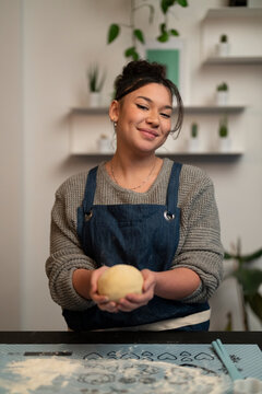 A young woman wearing an apron smiles while holding a ball of dough. She is preparing to bake in her kitchen, with flour visible on the counter.
