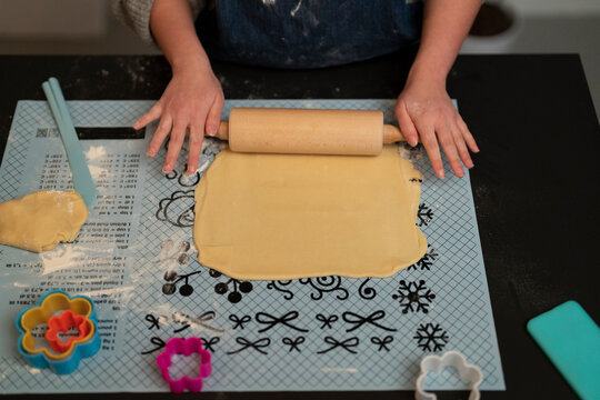 A person's hands are busy rolling out cookie dough on a patterned baking mat using a wooden rolling pin. Colorful cookie cutters sit nearby, ready for shaping the dough.