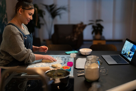 A young woman bakes at home, kneading dough on a mat. She follows a recipe tutorial on her laptop, surrounded by baking ingredients and tools. Enjoying the process of making homemade treats.