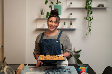 A young woman in an apron smiles while holding a tray of baked cookies. She is in her home kitchen, ready to share her sweet creations.
