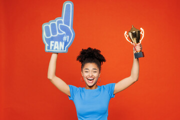 Young woman fan wears basic blue t-shirt foam 1 glove finger up cheer up support football sport team hold in hand golden cup soccer ball watch tv live stream isolated on plain red orange background.