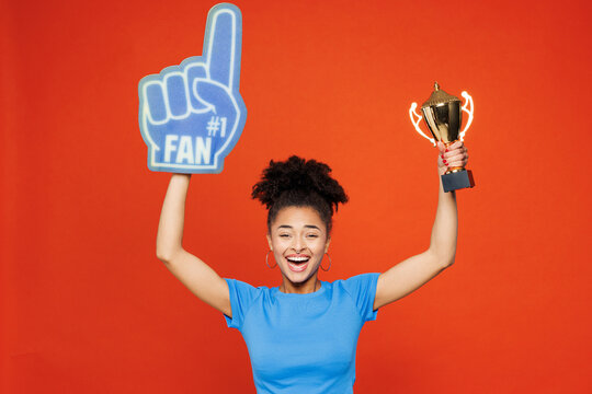 Young woman fan wears basic blue t-shirt foam 1 glove finger up cheer up support football sport team hold in hand golden cup soccer ball watch tv live stream isolated on plain red orange background. - Powered by Adobe