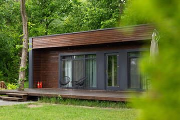 Modern wooden cabin with terrace in green forest, two chairs and fire extinguishers visible, peaceful summer retreat.