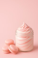 A jar of whipped body cream with strawberry frosting swirls and pastel pink macarons arranged in the lower left corner. At least two-thirds of the image is a soft, candy-colored background, left clean