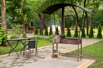 Outdoor barbecue grill with canopy, metal table, and chairs in a green garden setting, perfect for summer picnics and gatherings.