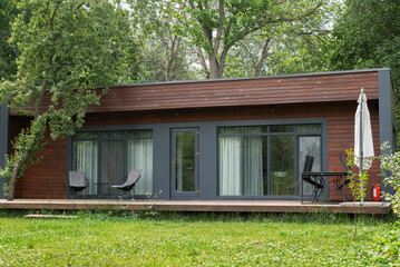 Modern wooden cabin with large windows, surrounded by greenery and trees, offering a peaceful retreat in nature.
