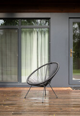 Modern black outdoor chair on a wooden terrace in front of a house with large windows and closed curtains.