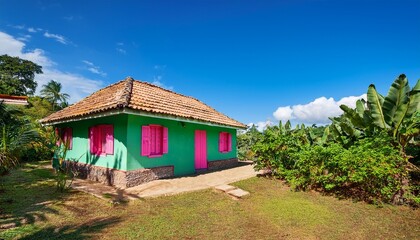Fototapeta premium small rural house with a vibrant green facade pink window shutters and a dark tiled roof surrounded by lush tropical vegetation under a clear blue sky