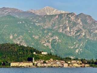 Scenic view from Griante and Cadenabbia villages on Lake Como shoreline with lush hills, alpine peaks and a ferry cruising on calm waters under a soft summer evening light