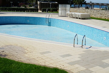 Empty tiled swimming pool in a resort with surrounding pavement and green grass, concept of off season, maintenance, renovation