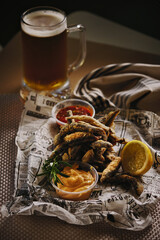 Close-up serving of crispy fried anchovies on vintage newspaper print, garnished with dipping sauces and glass of cold beer. Seafood, appetizers, Mediterranean cuisine.
