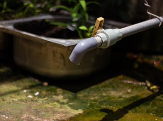 A grey water pipe with a yellow valve drips onto a mossy ground, next to an old sink with a plant, conveying themes of water conservation and DIY greywater recycling.


