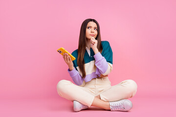 Charming Thoughtful young woman holding smartphone seated on pink background in casual outfit