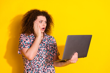 Young woman with curly hair and vibrant shirt looks shocked while using a laptop against a vivid...
