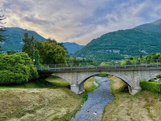 Old arched stone bridge between Lenno and Ossuccio, Italy, spanning a dry riverbed with green mountain backdrop and cloudy evening sky in a peaceful Lombard countryside setting