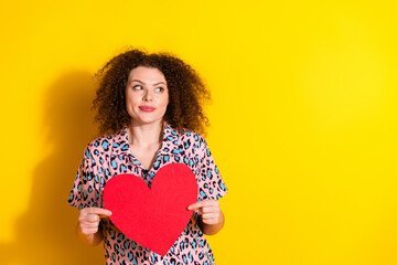 Young cheerful woman holding a red heart-shaped cutout on a vibrant yellow background, expressing...