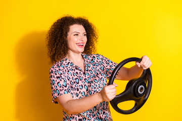 Joyful young woman holding a steering wheel against a vibrant yellow background, expressing...