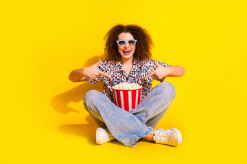 Joyful young woman in casual style pointing at popcorn against vibrant yellow background