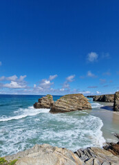 Obraz premium Scenic coastal view featuring rocky formations, gentle waves lapping at the shore, and a vibrant blue sky with scattered clouds, creating a serene beach atmosphere
