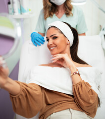 Beautiful and happy young woman sitting in medical chair and looking in the mirror. She is satisfied after successful beauty treatment with hyaluronic acid fillers or botulinum toxin injections.
