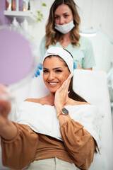 Beautiful and happy young woman sitting in medical chair and looking in the mirror. She is satisfied after successful beauty treatment with hyaluronic acid fillers or botulinum toxin injections.