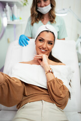 Beautiful and happy young woman sitting in medical chair and looking in the mirror. She is satisfied after successful beauty treatment with hyaluronic acid fillers or botulinum toxin injections.